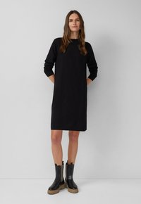 Frau, die mit den Händen in den Taschen steht, trägt ein schwarzes langärmliges knielanges Kleid und schwarze Stiefel mit klobigen, hellbraunen Sohlen.