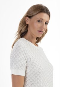 Hvid strikket sweater med et tekstureret skala-mønster, korte ærmer og rund hals. Materialet ser let og åndbart ud.