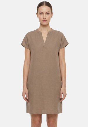 Donna che indossa un vestito taupe a maniche corte, lungo fino al ginocchio, con collo a V, in piedi su uno sfondo chiaro uniforme.