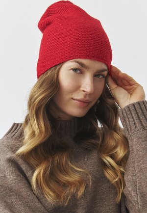 Beanie rosso lavorato a maglia con una vestibilità morbida. Motivo strutturato e design a cupola. Indossato con un maglione marrone a coste.