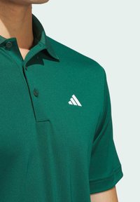 Vihreä polo-paita, joka on valmistettu sileästä kankaasta ja jossa on kauluspaita. Siinä on yksi nappi ja valkoinen Adidas-logo vasemmassa rinnassa.