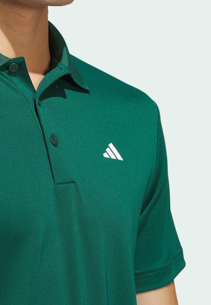 Grünes Polohemd aus glattem Stoff mit einem kragen. Besitzt einen einzelnen Knopf und ein weißes Adidas-Logo auf der linken Brust.