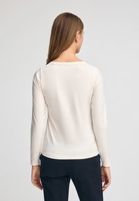 Langärmliges, cremefarbenes Hemd aus weichem Material. Verfügt über einen klassischen Rundhalsausschnitt und einen leicht geschwungenen Saum.