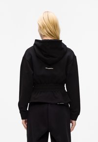 Sweat à capuche noir avec taille cintrée et détail de logo au dos, fabriqué dans un tissu doux et texturé. Dispose de manches longues.