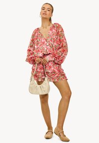 Vrouw in een roze bloemenjurk met pofmouwen, die een beige handtas vasthoudt, geaccessoireerd met gouden sieraden en beige flats.