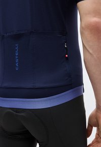 Gros plan sur une personne portant un maillot de cyclisme Castelli bleu marine avec une poche arrière zippée et un cuissard de cyclisme noir rembourré.