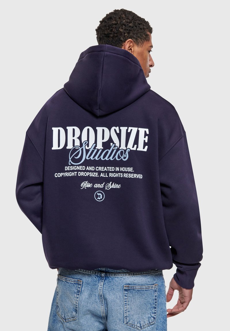Muškarac nosi mornarsko plavu majicu s kapuljačom s natpisom "Dropsize Studios" i dodatnim tekstom na leđima, u kombinaciji sa svijetloplavim trapericama, okrenut leđima prema kameri.