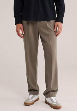 Homme portant un pantalon beige droit, des baskets blanches et beiges, et une chemise noire à manches longues, debout sur un fond neutre.