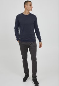 Marineblauer Pullover, Rundhalsausschnitt, lange Ä sleeves; kombiniert mit dunkelgrauen Hosen. Das Modell trägt schwarze Sneaker mit weißer Sohle. Glatte Textur, minimalistisches Design.
