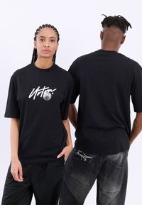 Svart bomull t-shirt med kort ärm, med en vit grafisk design och mån detaljer på framsidan. Bärs med svarta byxor och slitna jeans.