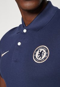 Donkerblauwe polo van textuurstof, met een wit logo van Chelsea Football Club en een kleine gouden Nike swoosh. Twee knopen zichtbaar.