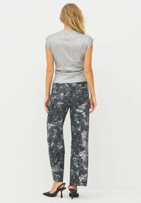 Žena stoji okrenuta leđima, noseći srebrni top bez rukava, tamno cvjetne hlače i crne sandale s visokom petom.