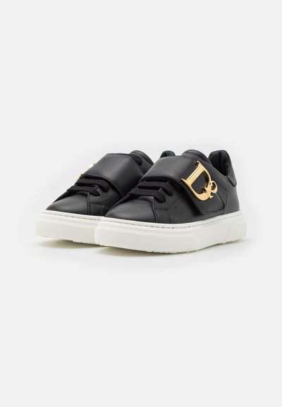 Sneaker in pelle nera con suola bianca. Presenta una larga vestibilità con un accento in oro del logo e lacci neri. Texture liscia e design minimalista.