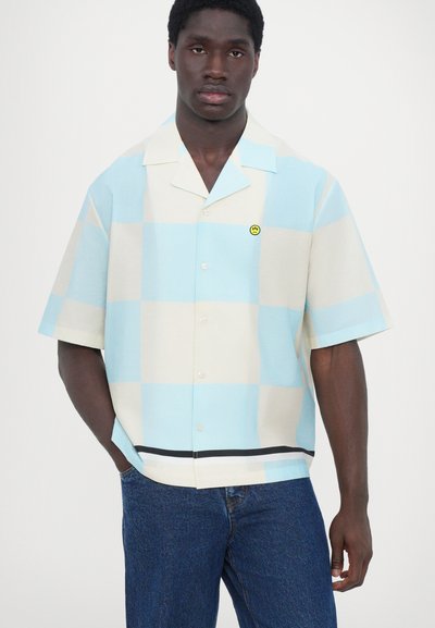 Camisa de manga corta hecha de una tela ligera, con un patrón a cuadros en azul y beige, cuello y un pequeño logo de cara sonriente amarilla.