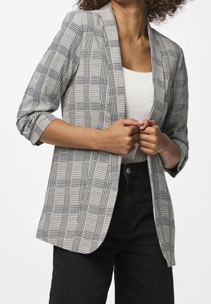 Personne portant un blazer à carreaux noir et blanc sur un haut blanc, associé à un pantalon noir taille haute, tenant les revers du blazer des deux mains.