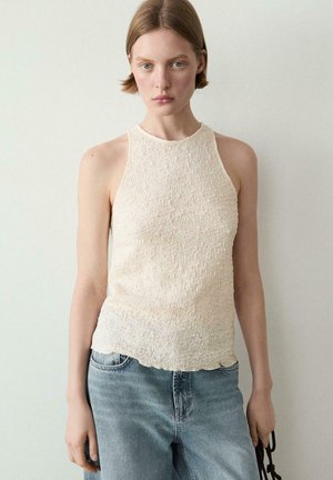 Jeune femme aux cheveux courts porte un haut crème texturé sans manches et un jean bleu clair, debout contre un mur clair uni.