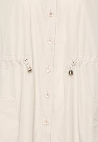Giacca button-up beige chiaro con vita elastica, due tasche laterali e cordini regolabili. Tessuto liscio e leggero con una finitura opaca.