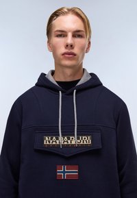Sudadera con capucha de color azul marino, con forro interior gris, que presenta un parche de logo prominente y una bandera noruega bordada en la parte frontal.