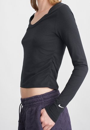 Femme portant un haut noir ajusté à manches longues avec fronces sur les côtés et pantalon sombre texturé avec cordon, main dans la poche.