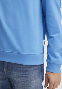 Sudadera azul claro con puños y dobladillo acanalados, con una etiqueta en el lateral. Lleva puesta con jeans azul oscuro, mostrando una textura de tela suave.