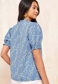 Blusa floreale blu con maniche corte a sbuffo, bordi arricciati e scollatura con arricciatura. Presenta un motivo di piccoli fiori bianchi e gialli.