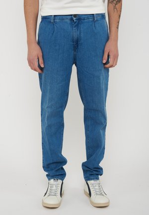 Uomo che indossa jeans blu a vestibilità rilassata e sneakers bianche con strisce nere, in piedi davanti a uno sfondo uniforme.