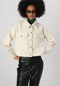 Cream-colored cropped jacket made of textured fabric, featuring a collared neckline, two front patch pockets, and snap closures.