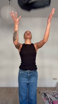 Osoba w czarnym topie na ramiączkach i niebieskich dżinsach rzucająca czarną kurtkę w górę w pomieszczeniu, na tle białej ściany i wiszącej lampy.