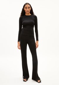 Schwarzes, geripptes Langarmoberteil, kombiniert mit taillierten schwarzen Hosen. Die Hosen haben seitliche Schlitze und sind mit schwarzen Sandalen mit quadratischen Zehen gestylt.