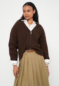 Pruuni kaabitsaga nööpidega kampsun, millel on valge kontrastne krae, koos volditud beeži seelikuga, millel on pruun vöö ja detailsed õmblused.