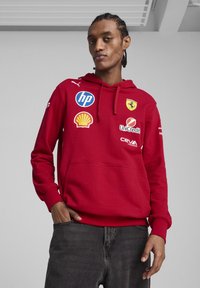 Sarkana kapuce džemperis, izgatavots no kokvilnas maisījuma. Iekļauj logotipus, tostarp Ferrari un Shell. Kenguru kabata un ribota piedurkne piešķir tekstūru.
