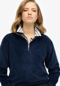 Sudadera de terciopelo azul marino con cuello alto, que presenta una cremallera y una camisa de rayas con cuello debajo. Logo sutil en el pecho.