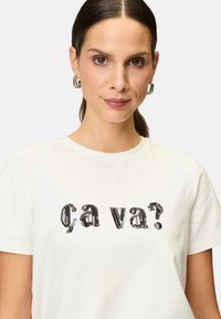 White cotton t-shirt featuring a black sequined text design "ça va?" in a stylized font. Short sleeves and a round neck. Soft texture.