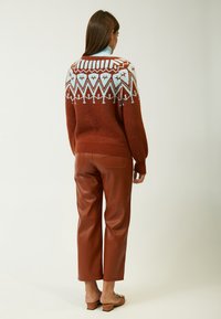 Ein rostfarbener Strickpullover mit einem gemusterten Schulterbereich in hellblau, kombiniert mit braunen Kunstlederhosen und beigefarbenen Slippern.