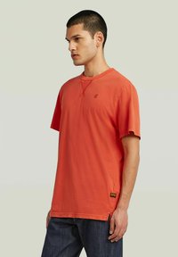 Camiseta de manga corta en coral brillante, hecha de algodón, con cuello redondo y pequeño detalle de logo en el pecho. Aberturas laterales en el dobladillo.