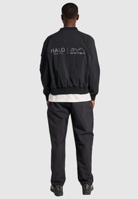 Czarna kurtka bombowca z gładkiego materiału, z elastycznymi mankietami i dołem, z białymi nadrukami logo na plecach. W połączeniu z ciemnymi spodniami.