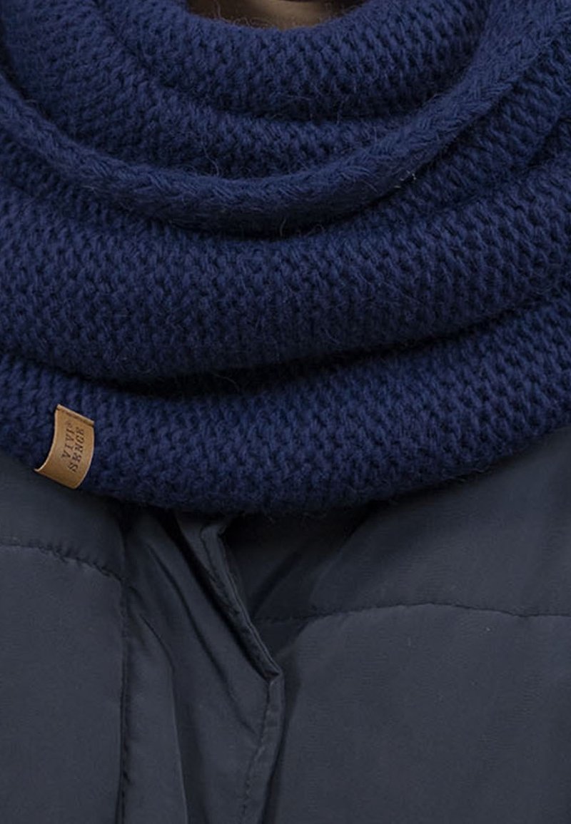 Écharpe épaisse en maille bleu marine enroulée autour du cou sur une veste matelassée foncée, avec une petite étiquette marron sur le bord de l’écharpe.
