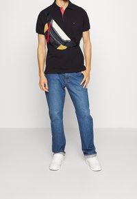 Schwarzes Polohemd mit rotem Kragen, blaue Jeans und weiße Sneaker. Eine schwarz-gelbe Umhängetasche mit einem Frontreißverschluss und roten Akzenten.