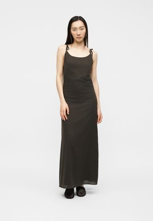 Kvinde iført en lang, mørk natkjole med tynde skulderstropper og sorte vævede slip-on sko, stående mod en ensfarvet hvid baggrund.