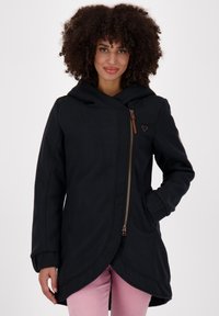 Donna con i capelli ricci che indossa un cappotto nero asimmetrico con zip e cappuccio e pantaloni rosa, in piedi con una mano in tasca.