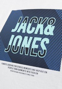 Valge T-särk, millel on erksate graafiliste tähtedega "JACK & JONES", helesinisel tumedate siniste triipudega taustal. Teksti üksikasjad on toodud allpool.