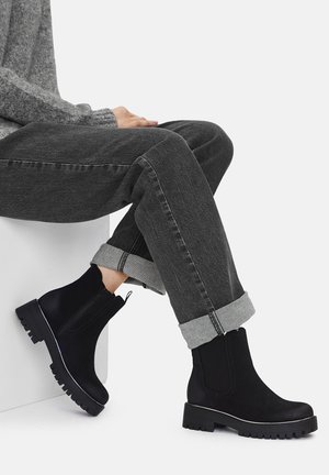 Czarne zamszowe botki za kostkę z grubą gumową podeszwą i bocznym zamkiem, zestawione z podwiniętymi szarymi dżinsami i szarym swetrem.
