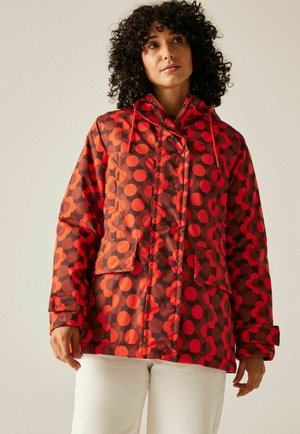 Kobieta o kręconych włosach, ubrana w czerwoną kurtkę w pomarańczowe grochy z kapturem oraz białe spodnie na jednolitym tle.