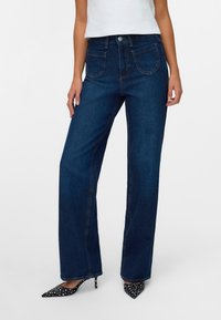 Vaqueros de pierna ancha de color azul oscuro con una cintura alta, diseño clásico de cinco bolsillos y costuras naranjas sutiles. Combinados con tacones negros de lunares puntiagudos.