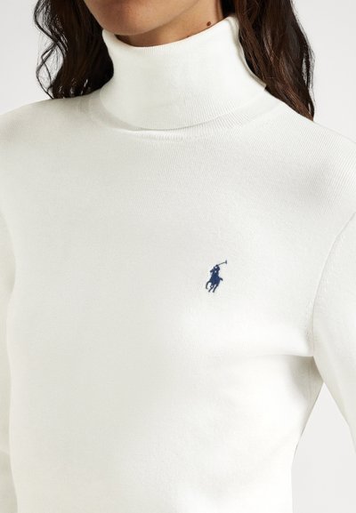 Žena nosi biely rebrovaný rolák s malým námornícko-modrým vyšívaným hráčom polo na ľavej strane hrudníka.