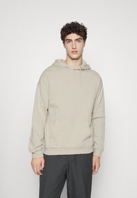Beige hoodie made of soft fabric, featuring a front pocket, adjustable drawstring hood, and relaxed fit. Paired with dark gray pants.