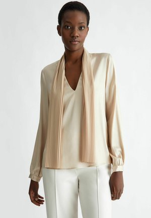 Blusa di satin beige con scollatura a V e maniche lunghe, caratterizzata da un dettaglio a sciarpa plissettata drappeggiata sul davanti, abbinata a pantaloni bianchi.
