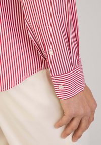 Rot-weißt gestreiftes Hemd mit langen Ärmeln, Knopfmanschetten, einem Umriss einer Brusttasche und glatter Stofftextur.