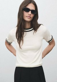 Woman with long brown hair wearing black oval sunglasses, white short-sleeve top with black trim, and black pants, standing against a plain background.