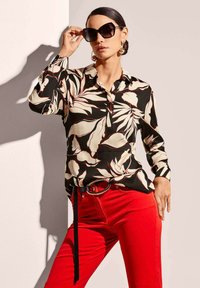 Long-sleeve shirt with black background and large white leaf pattern, paired with red slim-fit pants and a black belt, accessorized with oversized sunglasses.